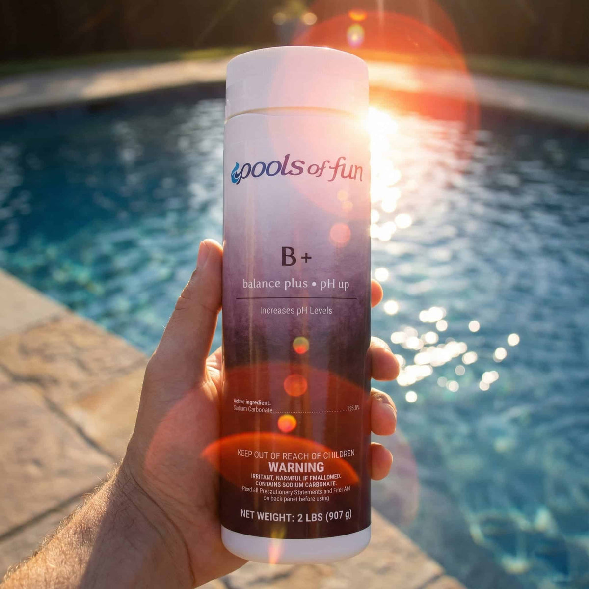 Hand holding a 'Pools of Fun' pool chemical bottle by a pool with sunset in the background